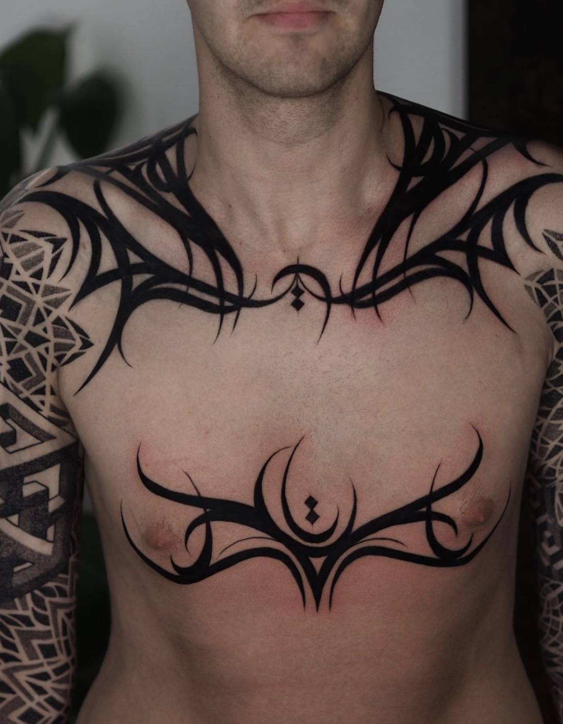 Man's chest tattoo featuring tribal-inspired black linework connecting across pectorals and shoulders
